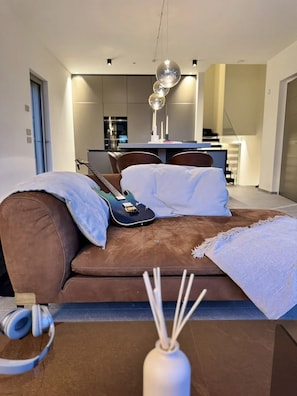 A modern bedroom with a brown sofa, a guitar, a white vase with reed sticks, and a dining area in the background. - Villa Aqua by Elite Villas (Tremezzina)