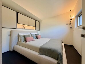 A bedroom with a bed, bedside tables, a wall-mounted light fixture, and a window. - Villa Aqua by Elite Villas (Tremezzina)