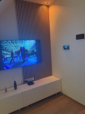 A modern living room with a large flat-screen TV mounted on a wall with vertical slats, a white entertainment unit below, and a small secondary screen on the wall. - Family home with a garden. Brussels is a 9-minute train ride away. Near Hallerbos (Halle)