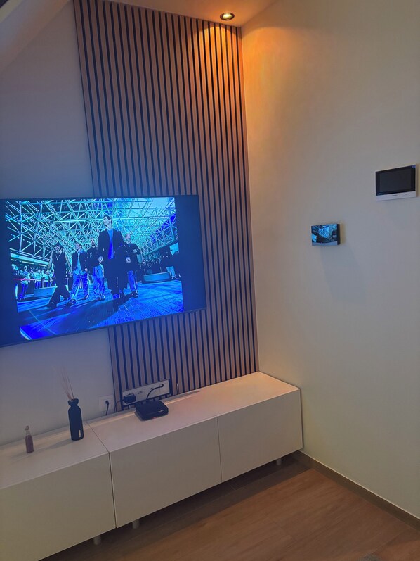 Ein modernes Wohnzimmer mit einem großen Flachbildfernseher an einer Wand mit vertikalen Lamellen, darunter ein weißes Unterhaltungselement und eine kleine, zusätzliche Anzeige an der Wand.