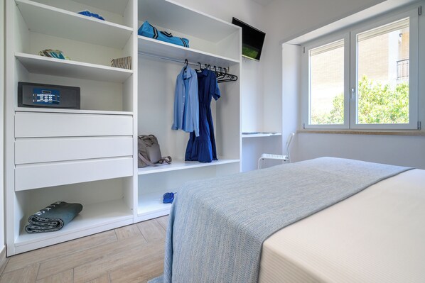 A bedroom with a bed, a wardrobe with hanging clothes, a television, and a window with blinds. - Casa Canzatora (Sperlonga)