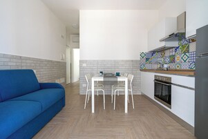 Un ambiente compatto con un divano blu, un tavolo da pranzo con sedie, un angolo cottura con fornello e forno, e uno schienale a parete decorato.