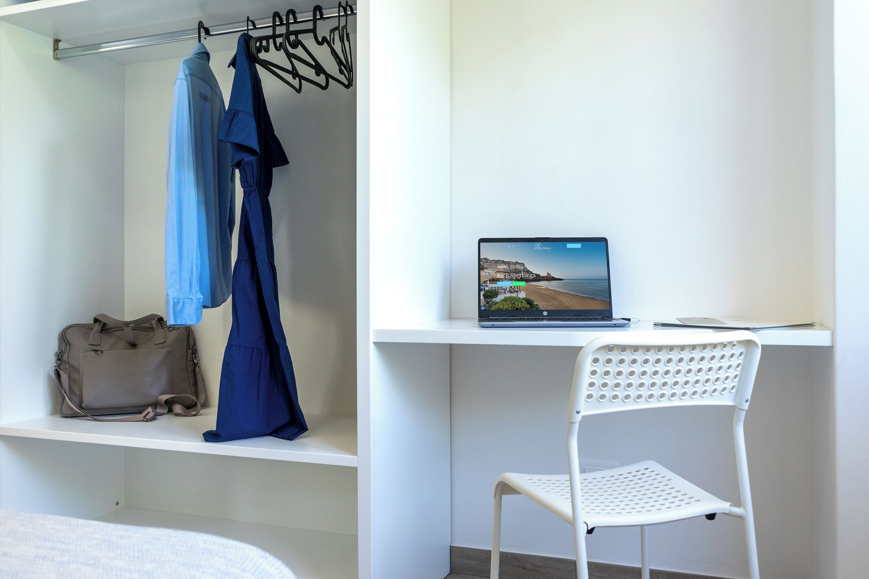 Una habitación con cama, un escritorio con una laptop que muestra una vista panorámica, una silla blanca y un perchero con un vestido azul.