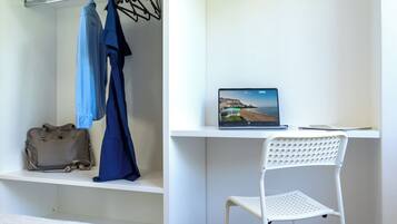 Una habitación con cama, un escritorio con una laptop que muestra una vista panorámica, una silla blanca y un perchero con un vestido azul.