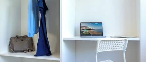 A room with a bed, a desk with a laptop displaying a scenic view, a white chair, and a clothing rack with a blue dress.