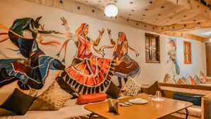 A room with a mural of dancers, a wooden table with a plate and glasses, and a bench with cushions.