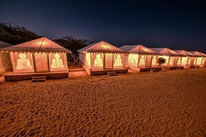 Una fila de tiendas de campaña con estatuas de Buda en la parte frontal, situadas en una playa arenosa de noche.