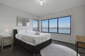 A bedroom with a large bed, a bedside table, a mirror, and a view of the sea through a large window. - Oceanview Oasis - Close to CBD and Airport (Sandy Bay)