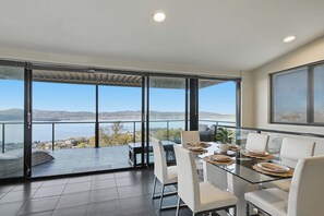 A modern dining area with a glass table and chairs, overlooking a scenic view of a body of water and distant hills. - Oceanview Oasis - Close to CBD and Airport (Sandy Bay)
