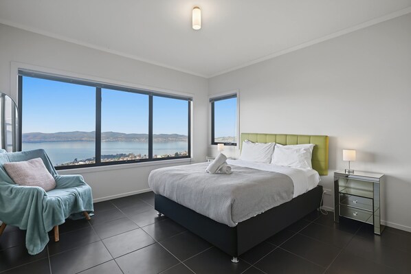 A modern bedroom with a large bed, a green headboard, and a view of the sea through large windows. - Oceanview Oasis - Close to CBD and Airport (Sandy Bay)