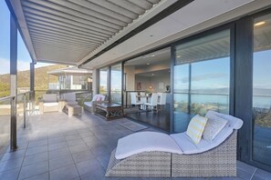 A modern outdoor living space with a glass-enclosed dining area, a lounge with a wicker sofa, and a view of the landscape. - Oceanview Oasis - Close to CBD and Airport (Sandy Bay)