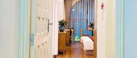 A hotel corridor with wooden flooring, a bed, a desk, and a chair.