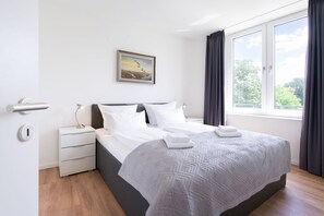 A modern bedroom with a large bed, a nightstand, a bedside table, a wall-mounted lamp, a window with curtains, and a framed picture on the wall. - Haus mit Spa-zugang in Meeresnahe (Lübeck)