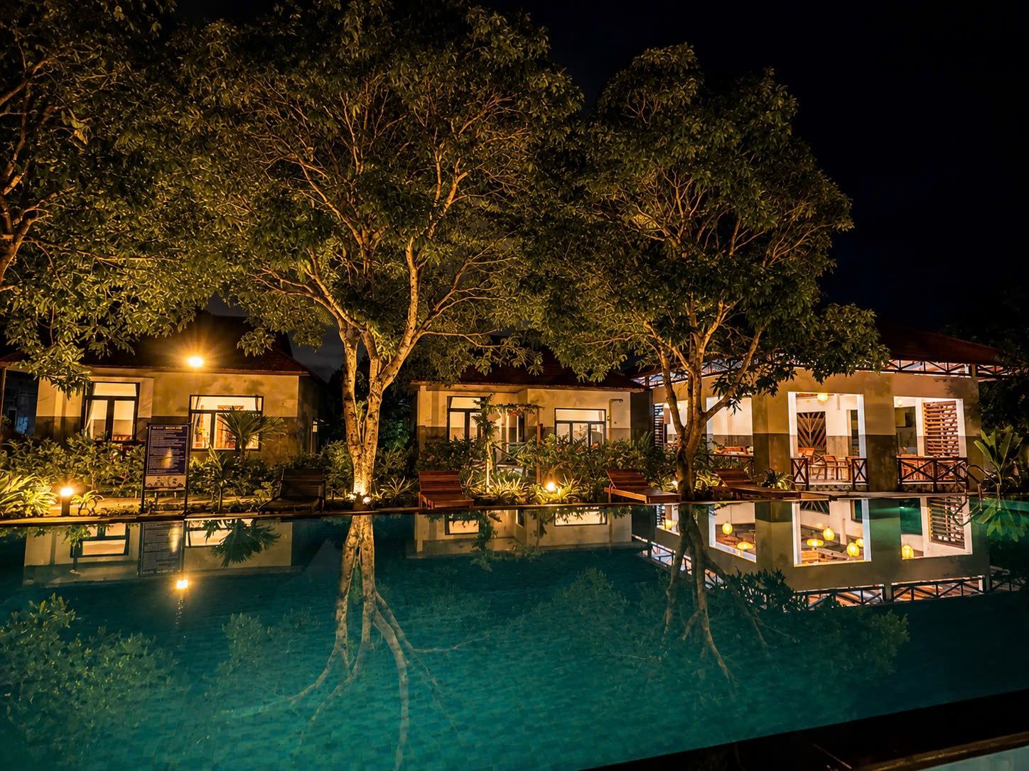 A nighttime view of a hotel with a swimming pool, lounge chairs, and trees.