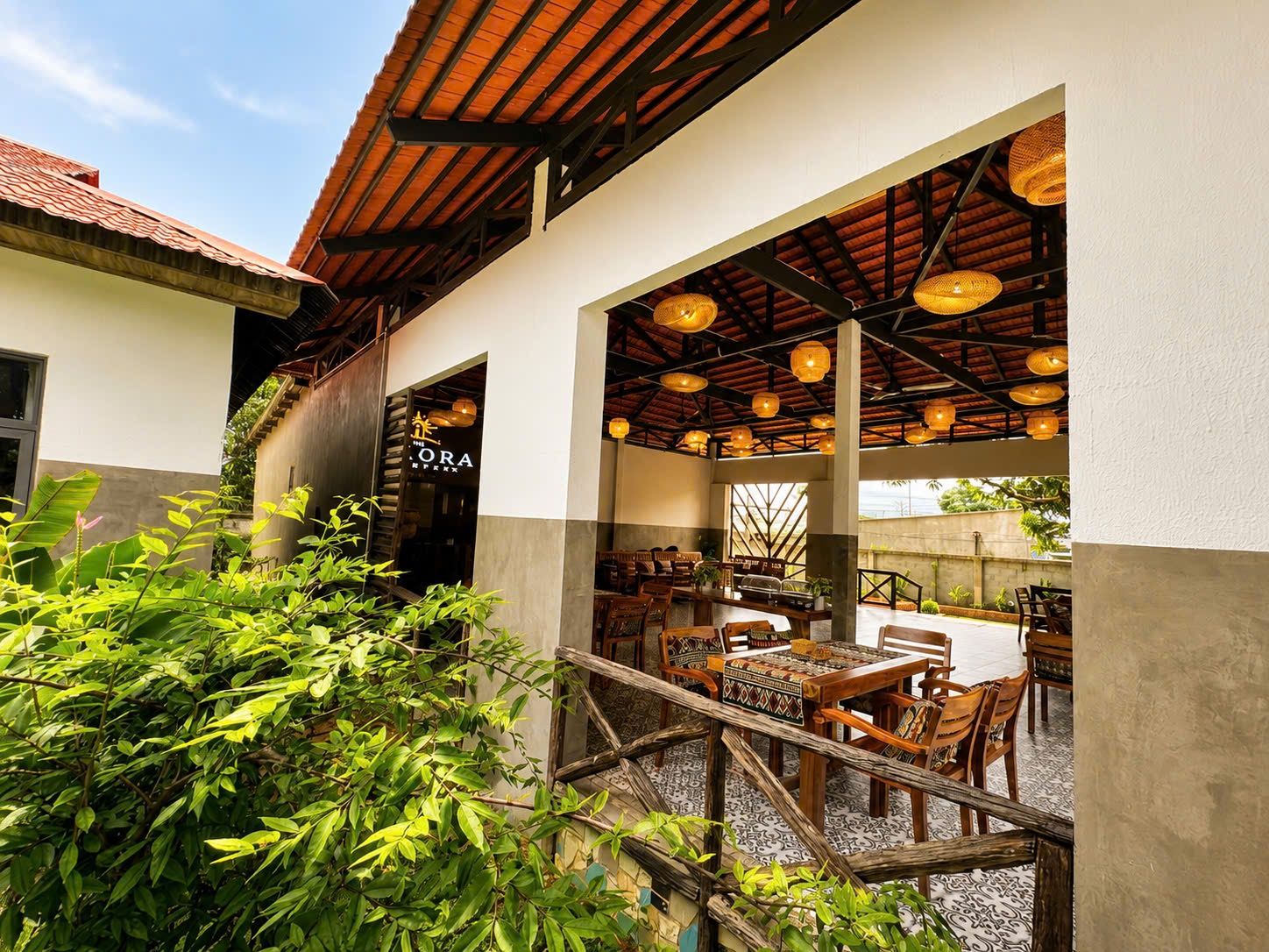 An outdoor dining area with tables and chairs, a covered seating area with hanging lights, and a building entrance with a sign.