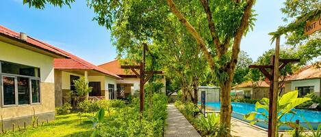 A pathway leading to a pool area with a building on the left and trees overhead.