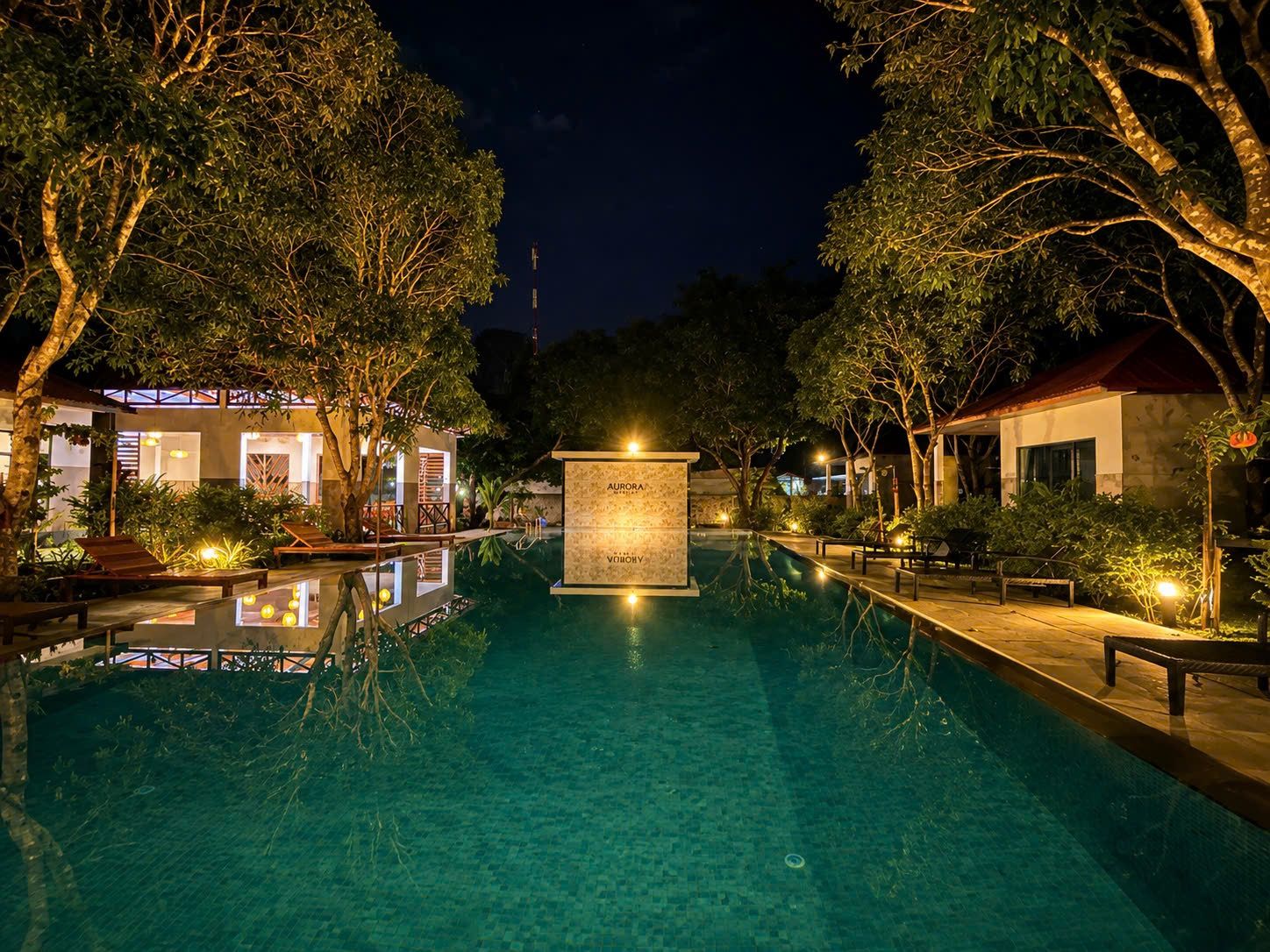 Una piscina nocturna con una fuente central, rodeada de árboles y edificios iluminados.