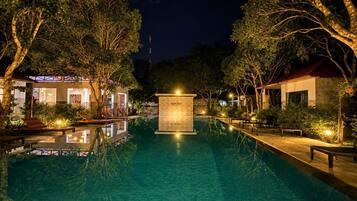 Una piscina nocturna con una fuente central, rodeada de árboles y edificios iluminados.