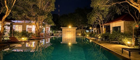 Piscina iluminada à noite, com uma fonte central, cercada por árvores e edifícios.