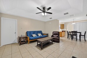 Un salon avec un ventilateur de plafond, un canapé, une table basse, une table à manger avec des chaises, et un coin cuisine à l’arrière-plan.