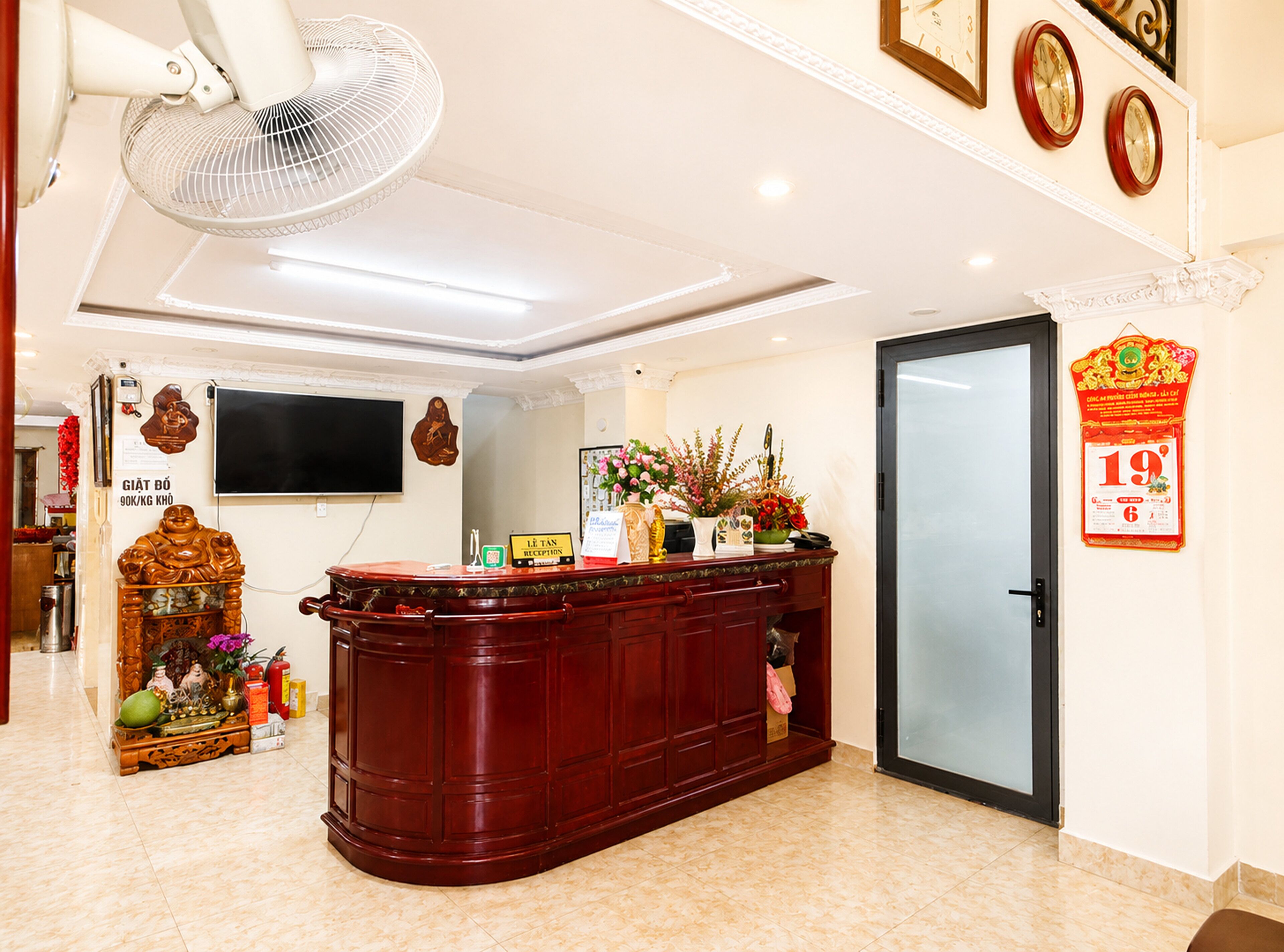A reception area with a wooden reception desk, a wall-mounted television, a clock, and a door with a red decoration.