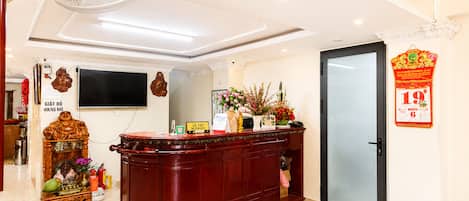 A reception area with a wooden reception desk, a wall-mounted television, a clock, and a door with a red decoration.