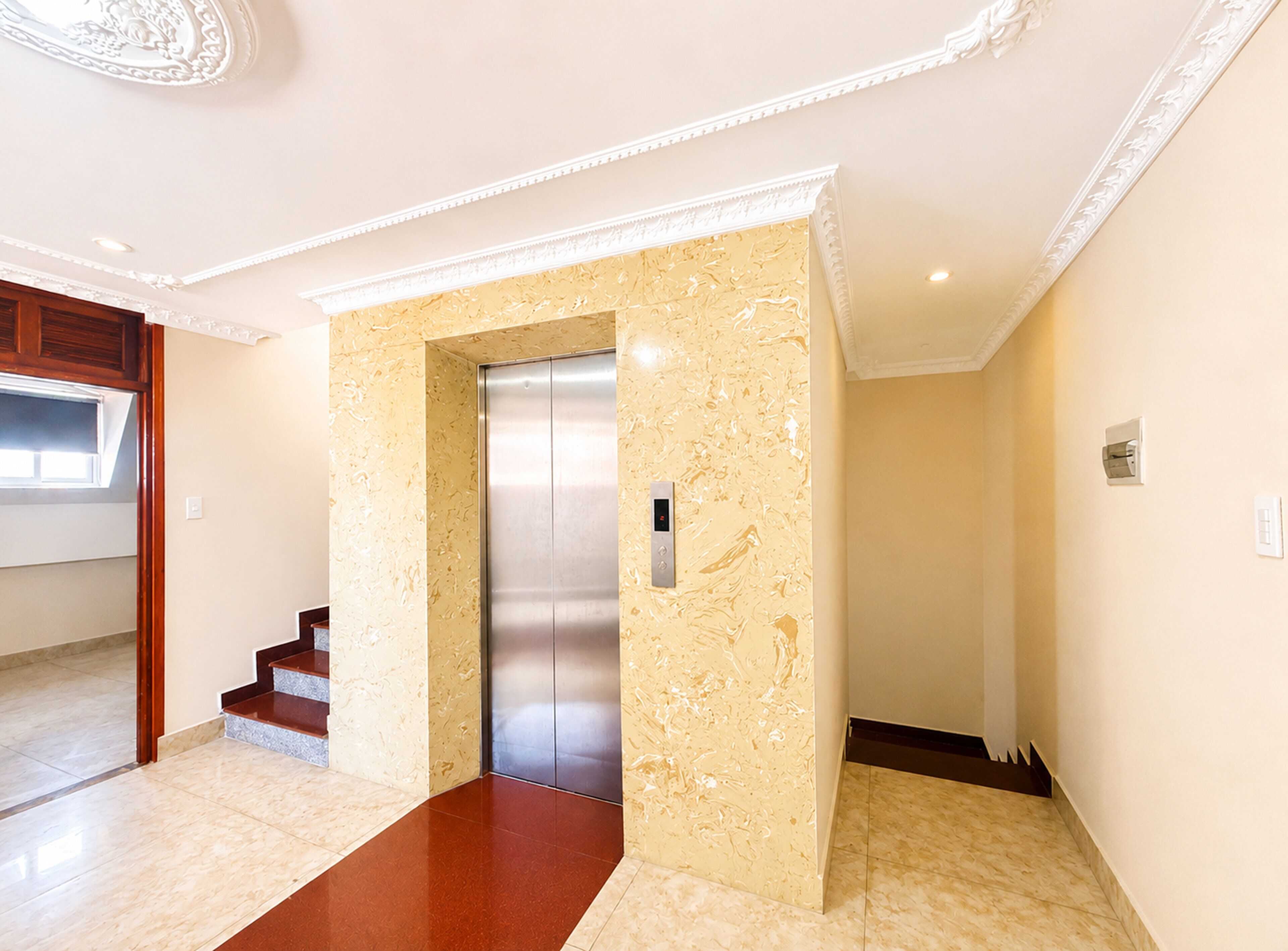 Un ascensor moderno con acabados metálicos, una pared de mármol con textura y una escalera con escalones de madera.