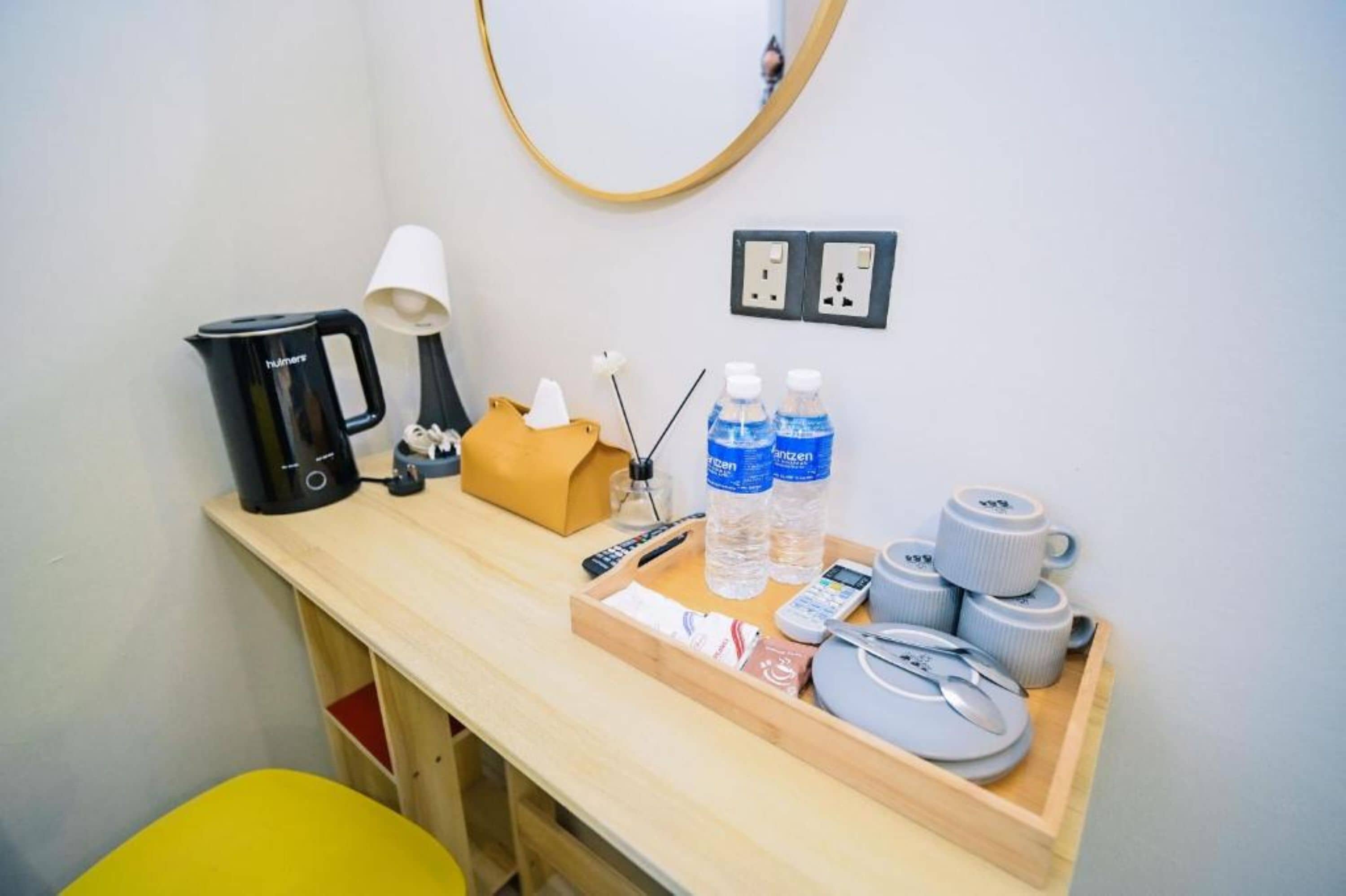 A hotel room with a wooden desk, a coffee maker, a lamp, a tissue box, bottled water, a remote control, a tray with a plate, and a pair of scissors.