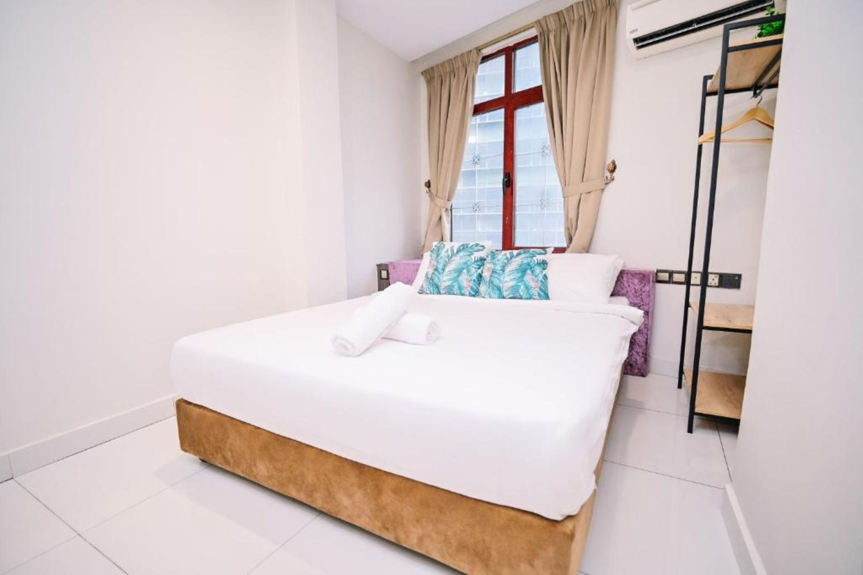 A neatly made bed with white bedding and a brown headboard, a window with beige curtains, and a wardrobe with a black frame.