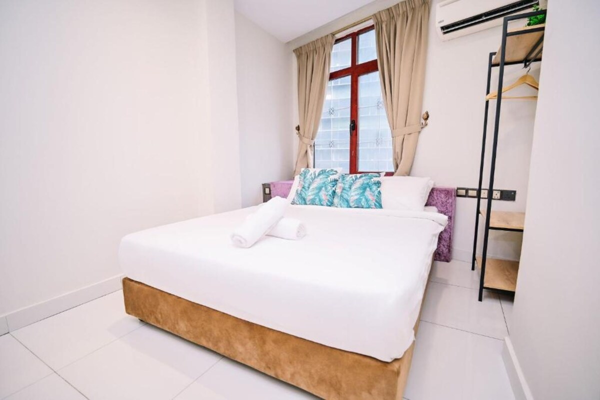 A neatly made bed with white bedding and a brown headboard, a window with beige curtains, and a wardrobe with a black frame.