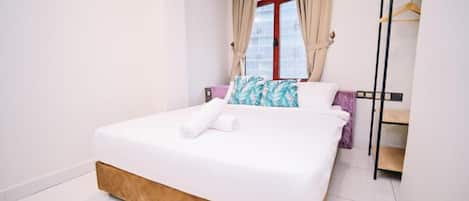 A neatly made bed with white bedding and a brown headboard, a window with beige curtains, and a wardrobe with a black frame.