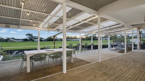 Een overdekt terras buiten, met een houten terras, metalen stoelen, een tafel en uitzicht op een grasveld.