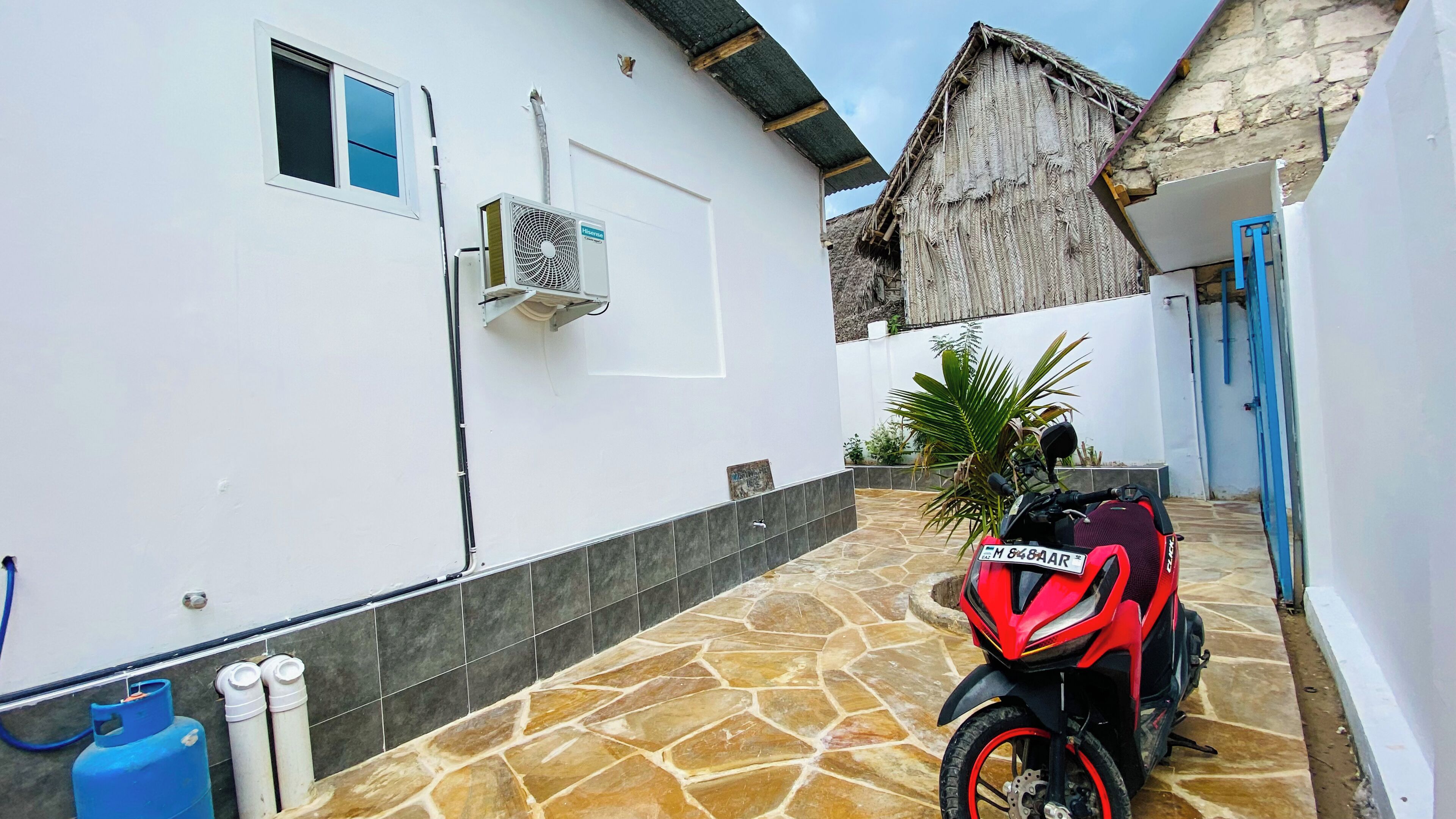 Una motocicleta estacionada en un área techada con piso de baldosas de piedra, un edificio blanco con ventana y unidad de aire acondicionado, y una estructura con techo de paja al fondo.