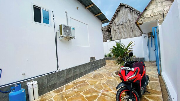 Een motorfiets geparkeerd in een overdekte ruimte met een vloer van stenen tegels, een wit gebouw met een raam en een airconditioner, en een gebouw met een rieten dak op de achtergrond.