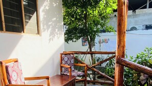 Een houten terras met een bank en stoelen, een betegelde vloer, en uitzicht op een boom en gebouw op de achtergrond.