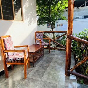 Un espace extérieur aménagé avec des meubles en bois, comprenant un banc et des chaises, un sol carrelé et une vue sur un arbre et un bâtiment en arrière-plan.