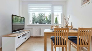 A modern dining area with a wooden table and chairs, a television on a white cabinet, and a view of trees through the windows. - Cosy Apartment for 6 People | Balcony | Opera Leśna (Sopot)