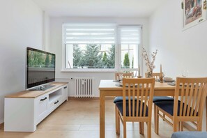 Ein moderner Essbereich mit einem Holztisch und Stühlen, einem Fernseher auf einem weißen Schrank und Blick auf Bäume durch die Fenster.