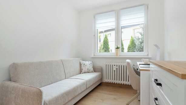 A minimalist room with a sofa, a radiator, and a window with a view of trees and buildings. - Cosy Apartment for 6 People | Balcony | Opera Leśna (Sopot)