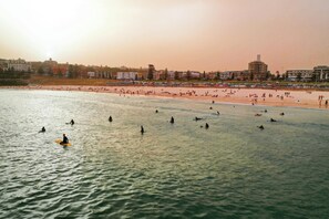 Ein Strand mit surfenden und schwimmenden Personen, Gebäuden im Hintergrund und einem klaren Himmel.