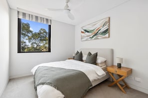 A bedroom with a bed, a nightstand, a lamp, a ceiling fan, a window with a view of trees, and a painting on the wall. - Modern 2BR Apartment within Prestigious Bellevue Hill (Bellevue Hill)