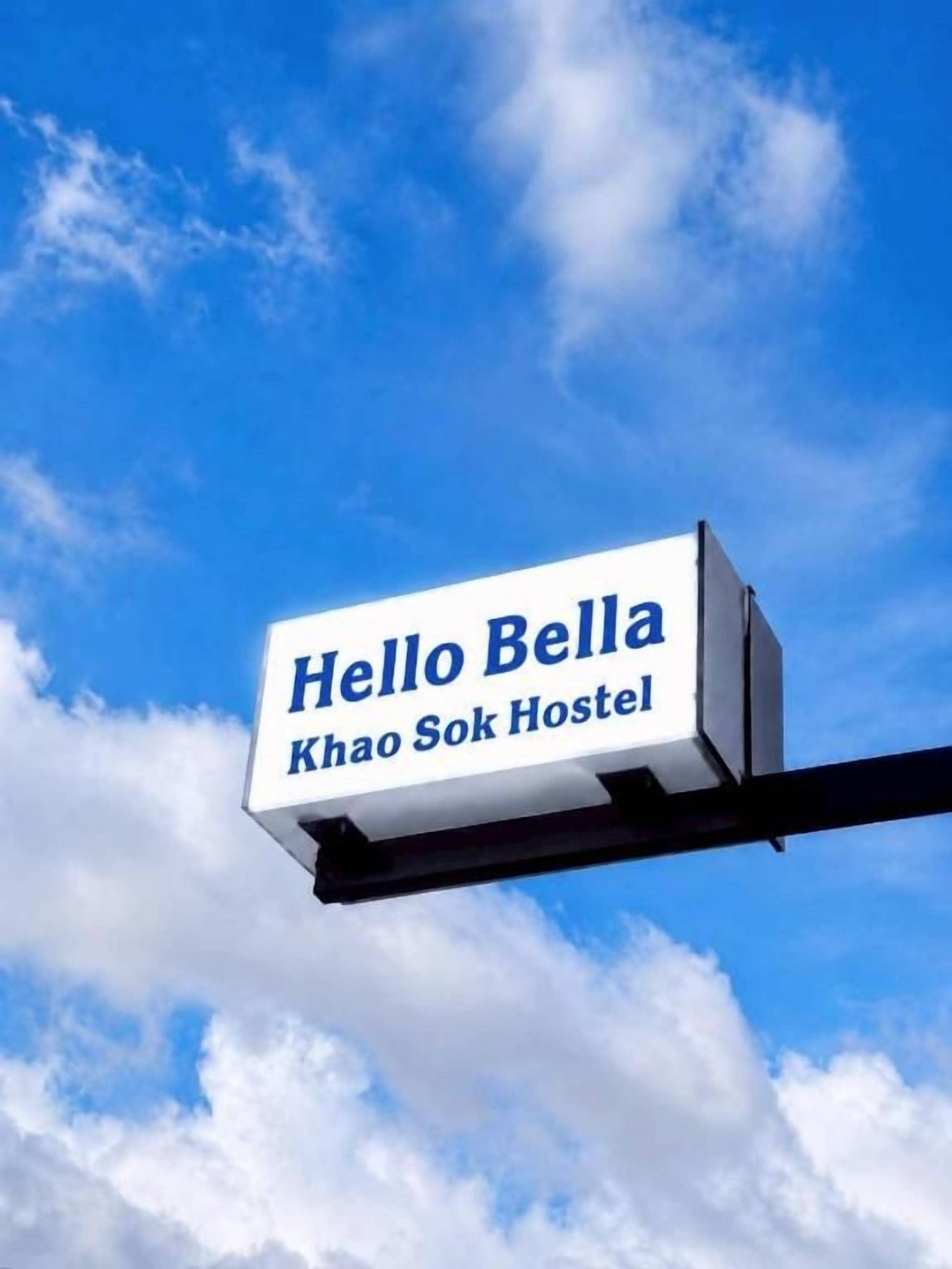 Un letrero con el texto 'Hello Bella' y 'Khao Sok Hostel' sobre un cielo azul con nubes.