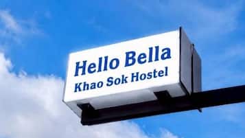 Un letrero con el texto 'Hello Bella' y 'Khao Sok Hostel' sobre un cielo azul con nubes.
