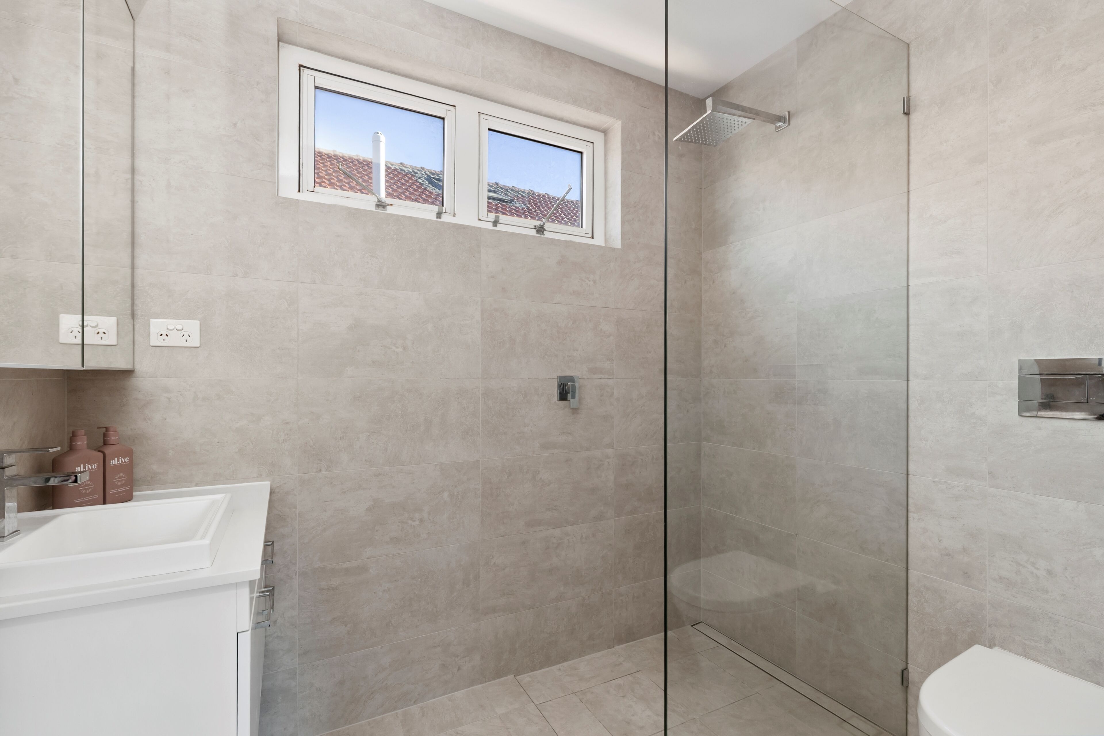 A modern bathroom with a glass-enclosed shower, a white sink, and a window above the shower.
