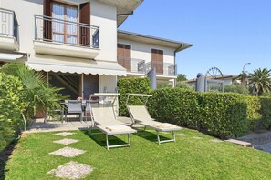 A two-story building with a balcony, a covered outdoor seating area with chairs and a table, and a well-maintained garden with hedges and a lawn. - Regarda - Basiliko View Apartment with lake view and pool (Lazise)