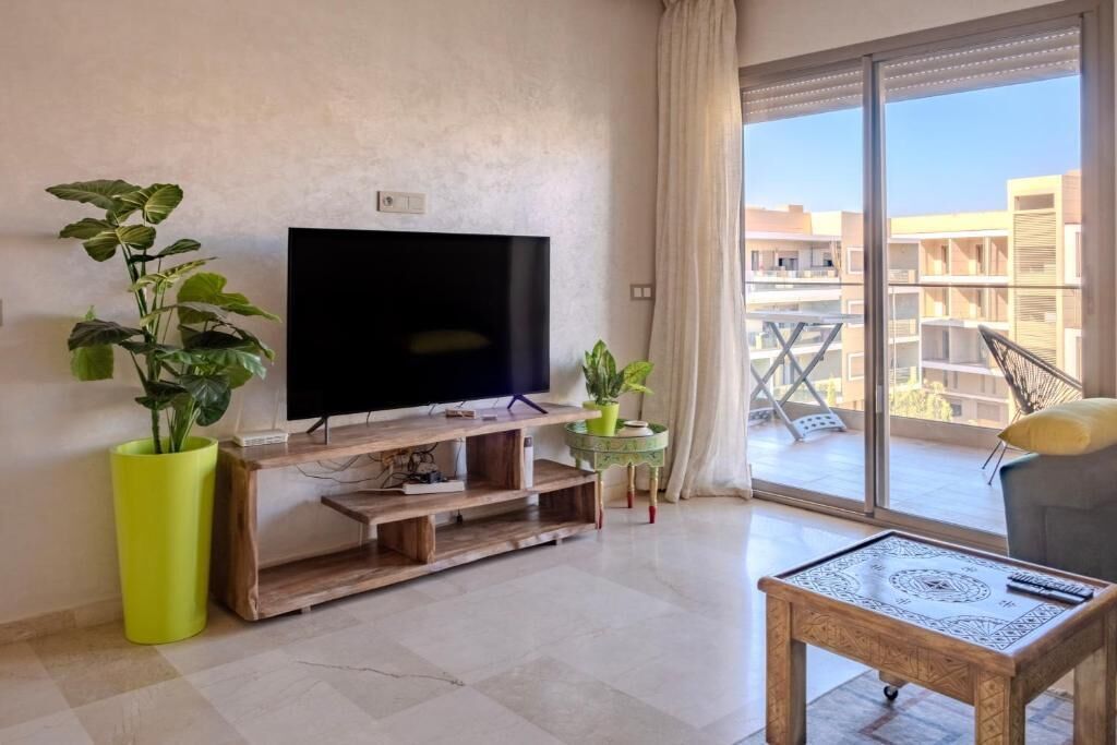 Un salon doté d’un téléviseur à écran plat, d’un meuble de rangement en bois, de plantes en pot, d’un balcon offrant une vue sur les bâtiments et d’une table basse.