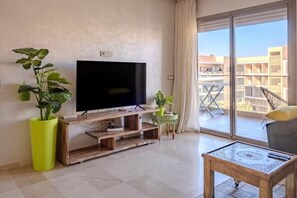 A living room with a flat-screen TV, wooden entertainment unit, potted plants, a balcony with a view of buildings, and a coffee table. - Oasis Pearl Garden luxe et confort (Marrakech)