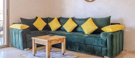 Um sofá verde com almofadas amarelas, uma mesa de centro de madeira e um tapete estampado.