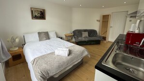 Habitación de hotel con cama, mesita de noche, ventilador y una kitchenette con lavabo y cafetera.