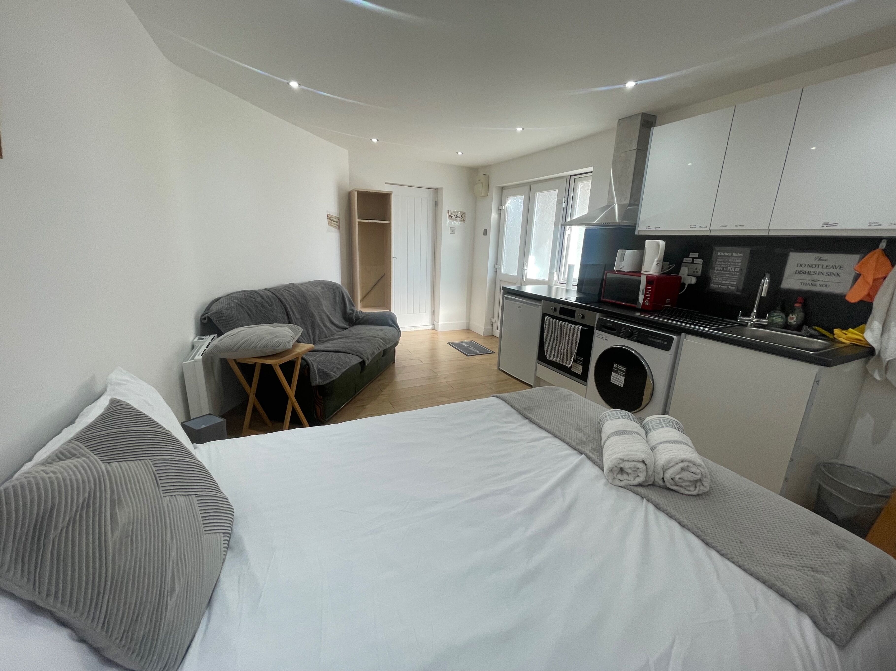 A compact hotel room with a bed, a sofa, a small table, a washing machine, and a kitchenette.